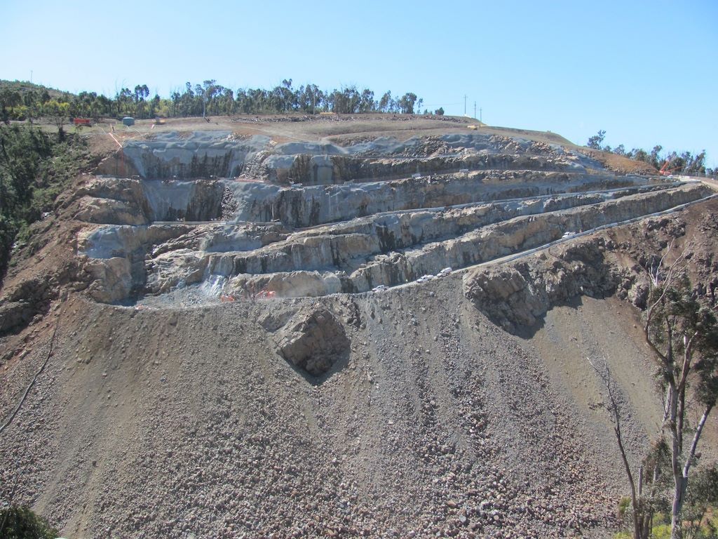 A terraced quarry on a hillside, showing exposed rock layers and scattered loose stones. Sparse trees surround the quarry, and a clear blue sky is visible above.