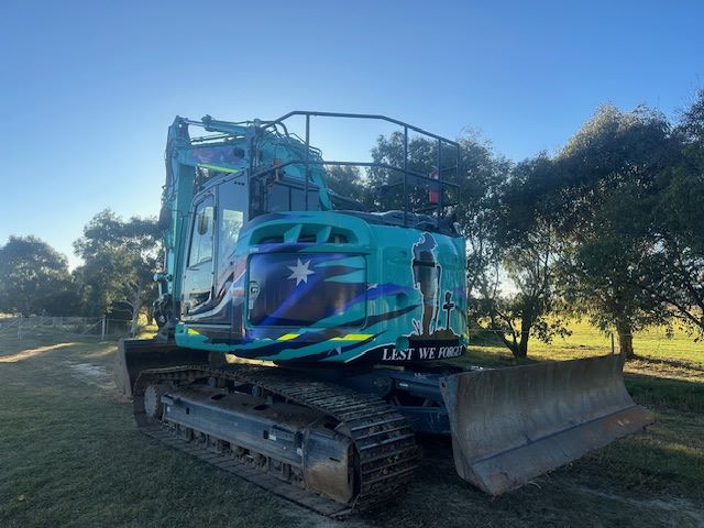 A blue excavator with military-themed artwork, including a soldier silhouette and the text "Lest We Forget," sits on a dirt path surrounded by trees under a clear blue sky.