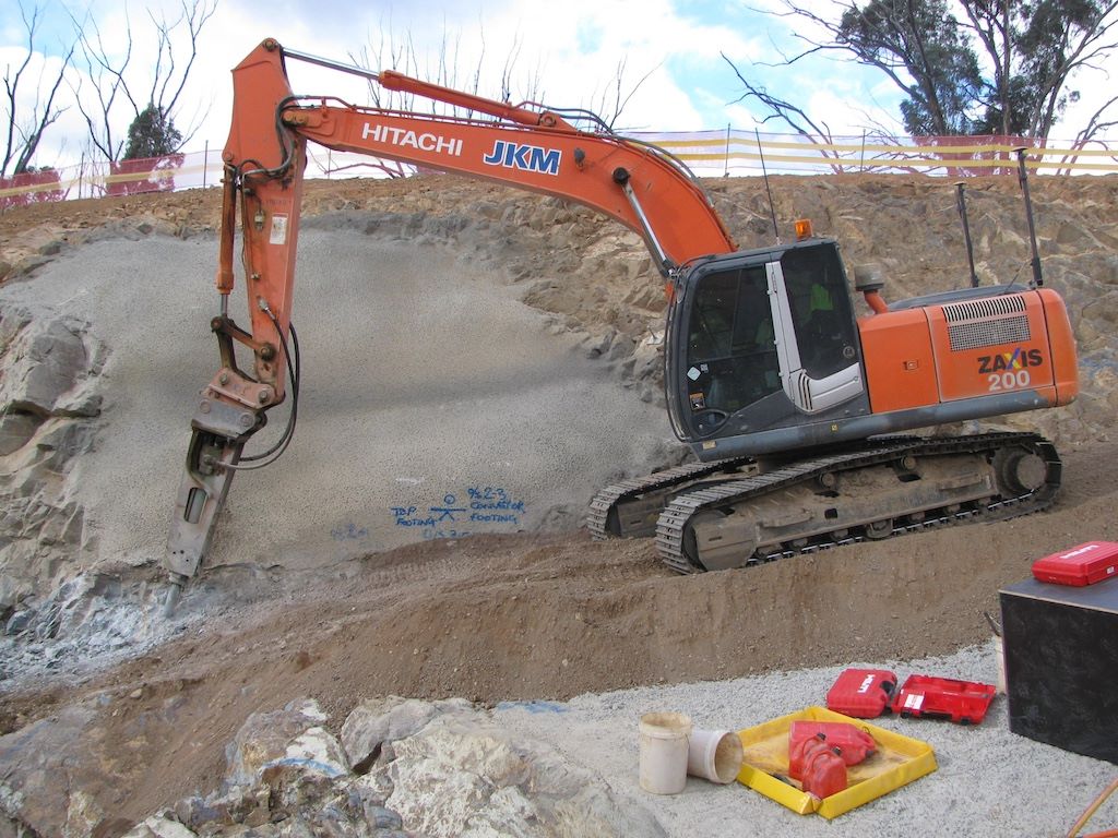 An orange excavator with a hydraulic breaker attachment is working on rocky terrain, breaking down a hillside. Equipment and tools are scattered in the foreground. Sparse, burned trees are visible in the background.