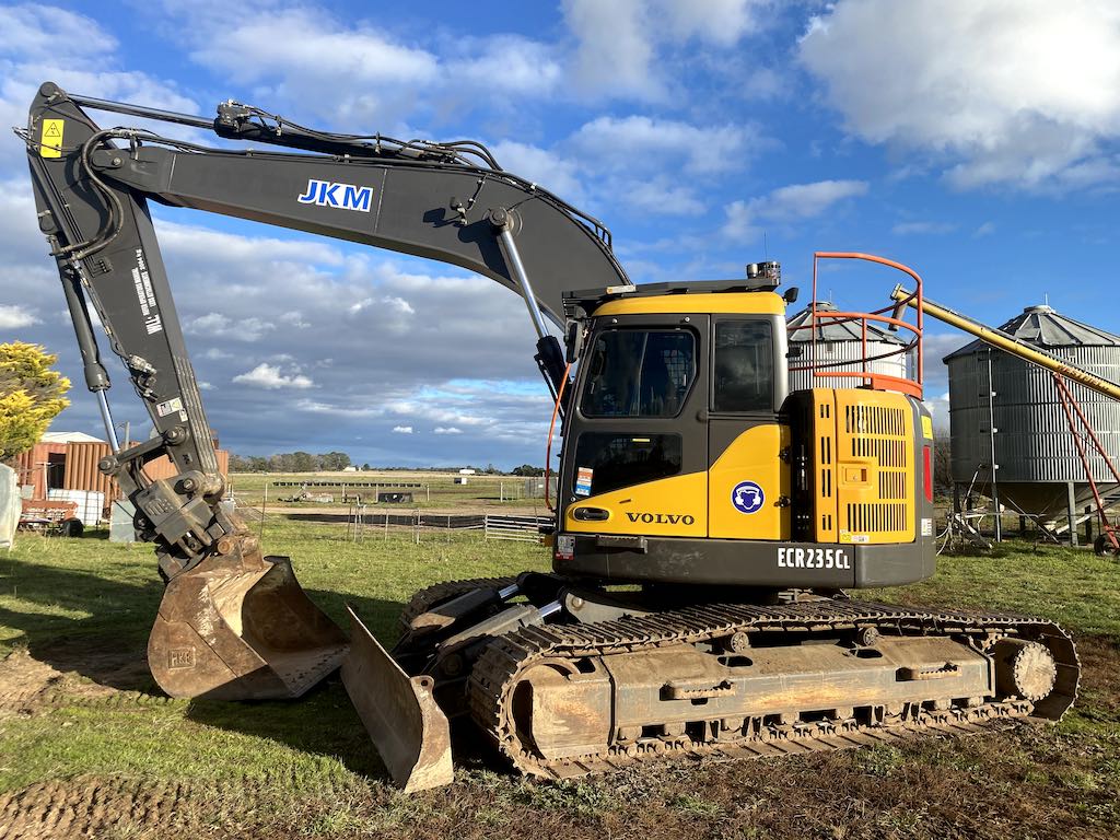 A yellow and black excavator with the label "JKM" on the arm is parked on grassy terrain. The sky is partly cloudy, and a silo is visible in the background.