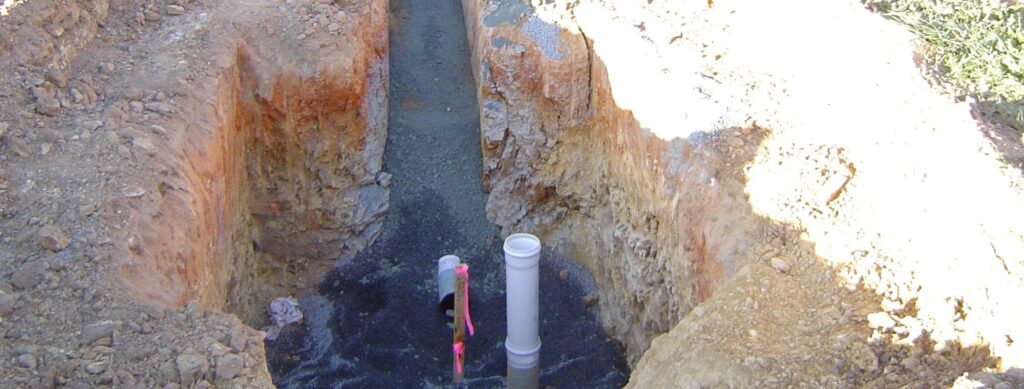 A trench with a partially buried PVC pipe and a smaller pink-marked pipe. The surrounding dirt is reddish-brown, and the trench is filled with gravel at the bottom. Sunlight casts shadows on one side of the trench.