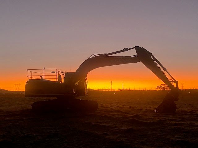 Silhouette of an excavator against a vibrant orange and pink sunset, with power lines and a few trees visible on the horizon. The scene conveys a peaceful yet industrial atmosphere.