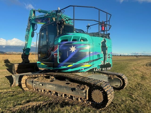 A teal excavator with a custom design features a flag, a silhouette of a soldier, and the phrase
