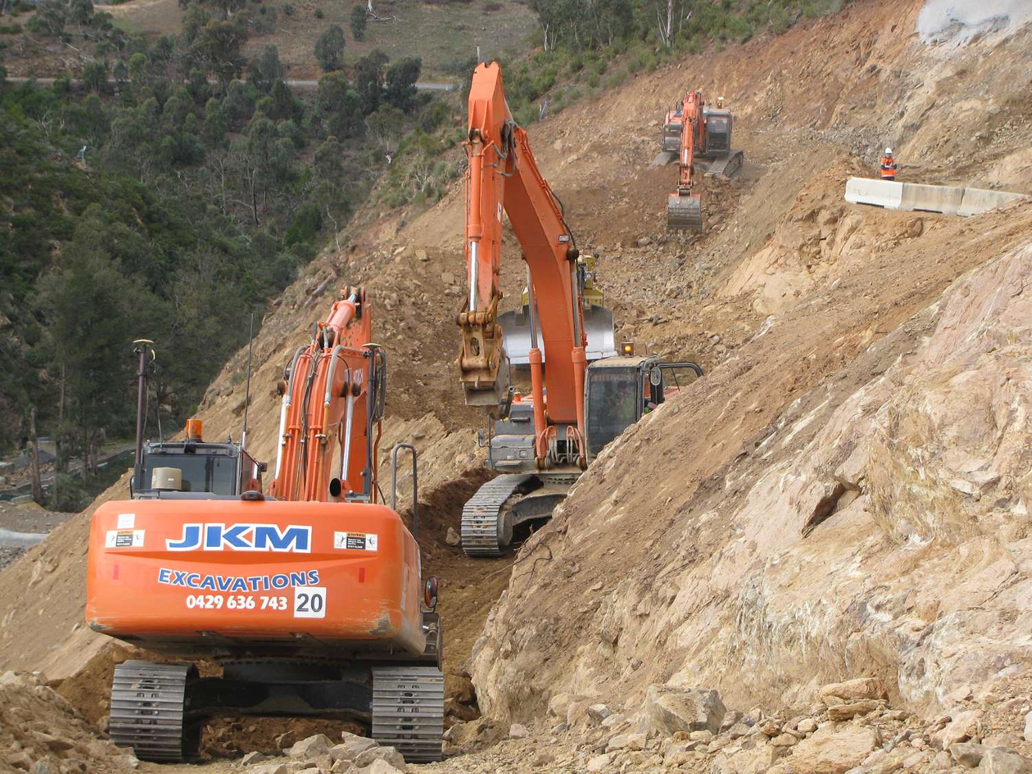 Three orange excavators work on a steep, rocky hillside, each placed at different heights. A construction worker in safety gear stands on the upper right side. The background features trees and a distant view of the landscape.